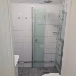 Nowoczesna łazienka z białymi, kwadratowymi płytkami, szklanym prysznicem z deszczownicą i półkami, widoczny fragment umywalki i toalety, szare płytki podłogowe.