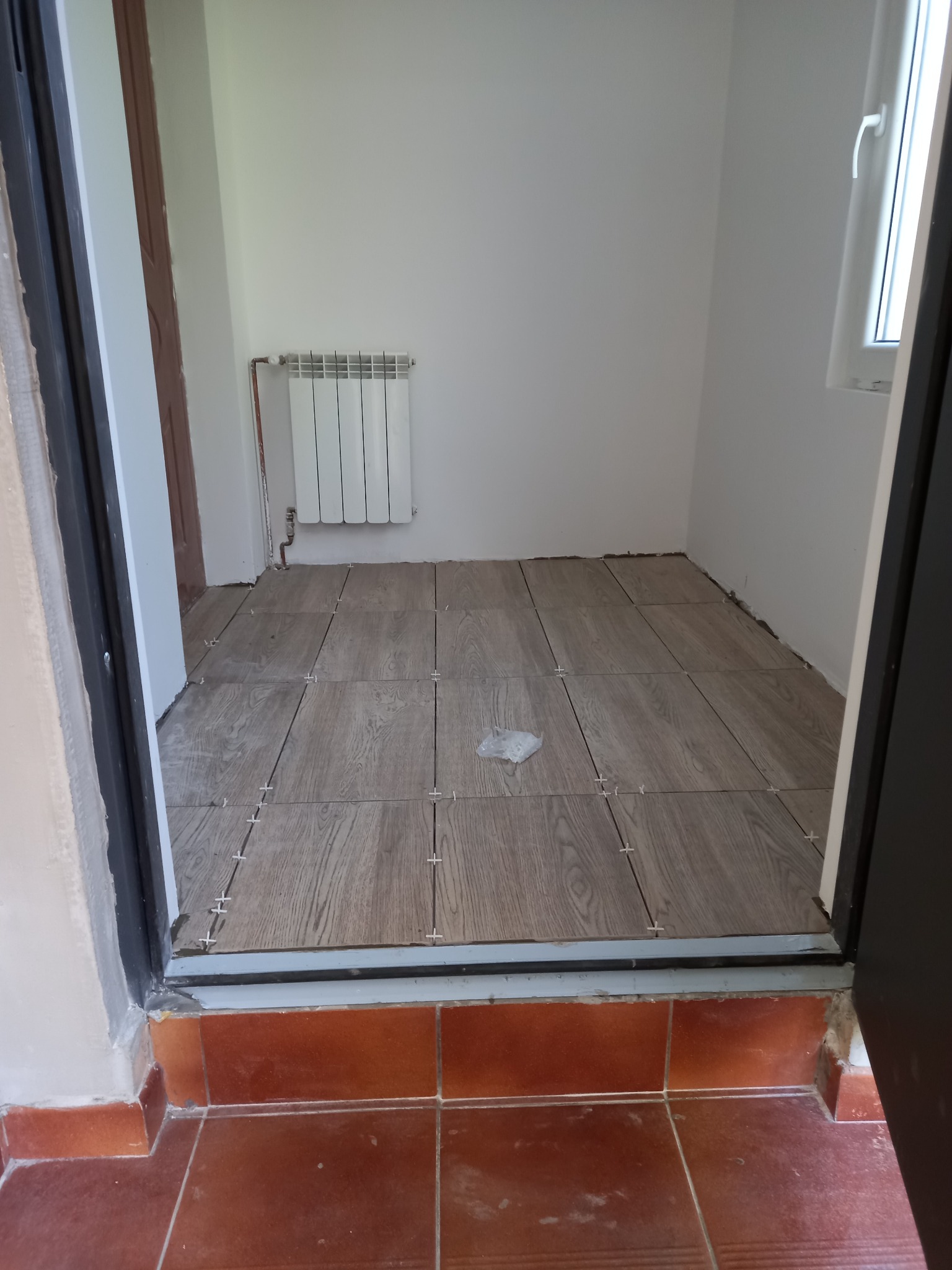 Układanie płytek imitujących drewno na podłodze w pomieszczeniu, widoczne krzyżyki dystansowe, grzejnik na ścianie, widok z progu.