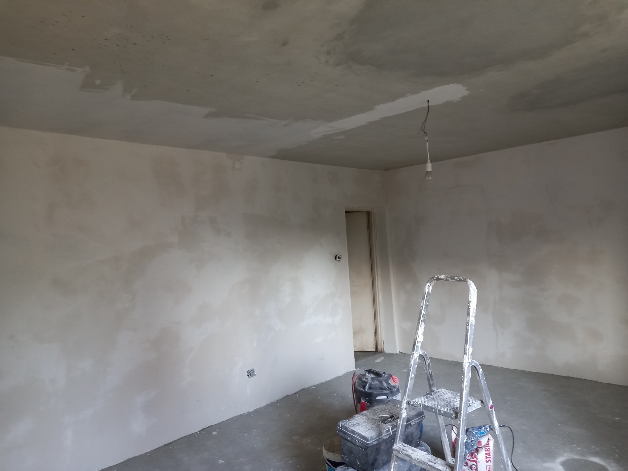 Pokój w trakcie prac wykończeniowych, ściany i sufit po nałożeniu gładzi, widoczne narzędzia, drabina i lampa wisząca na kablu.