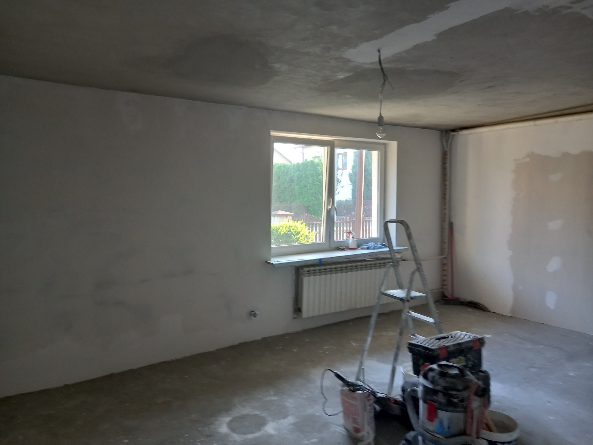 Pokój w trakcie remontu: ściany i sufit po szpachlowaniu, widoczna drabina, narzędzia i wiadro z zaprawą, okno z widokiem na zieleń.