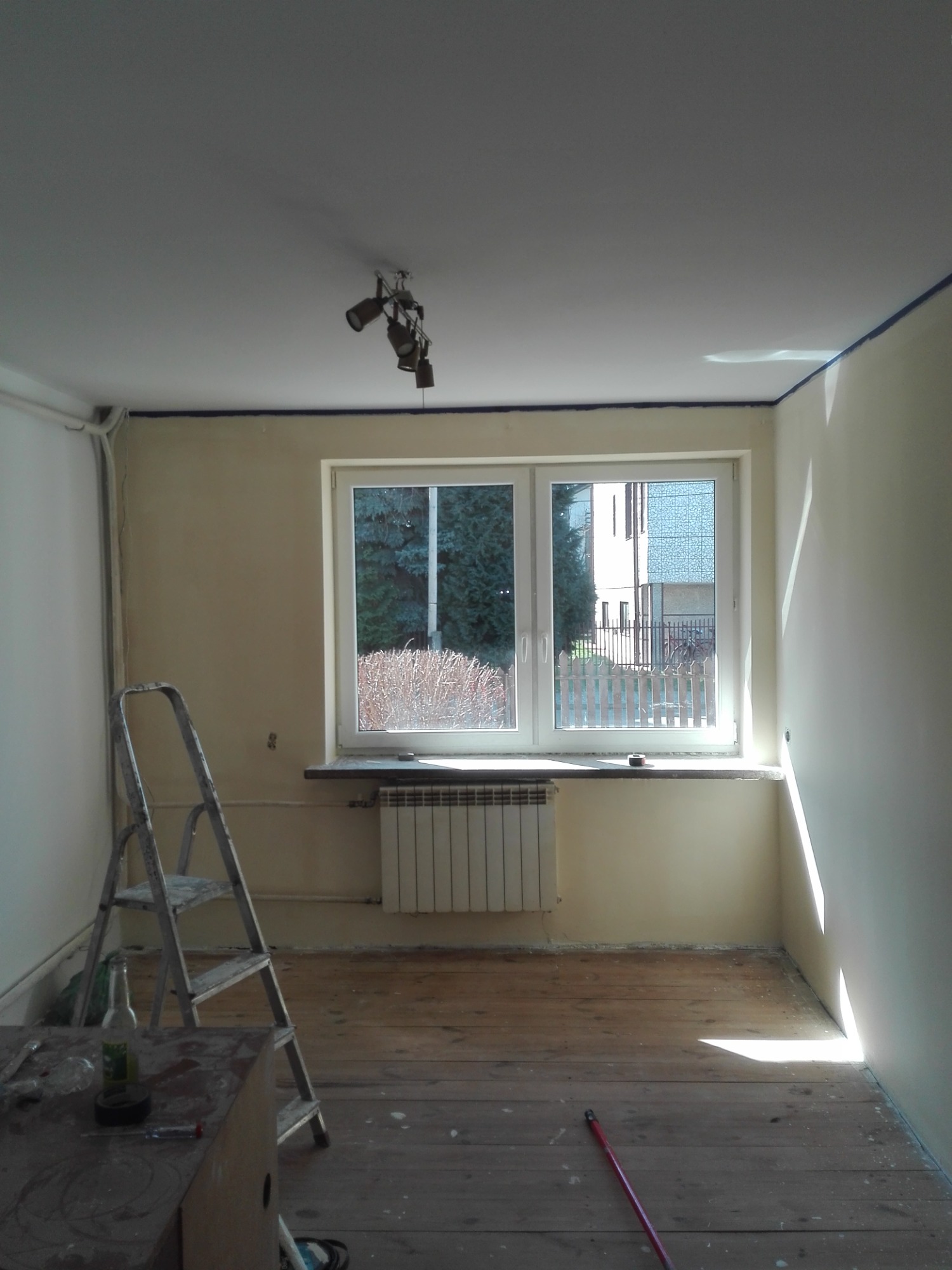 Pokój w trakcie przygotowania do malowania: ściany zagruntowane, zabezpieczone taśmą malarską przy suficie, drewniana podłoga częściowo zakryta, drabina oparta o ścianę, widoczne narzędzia...