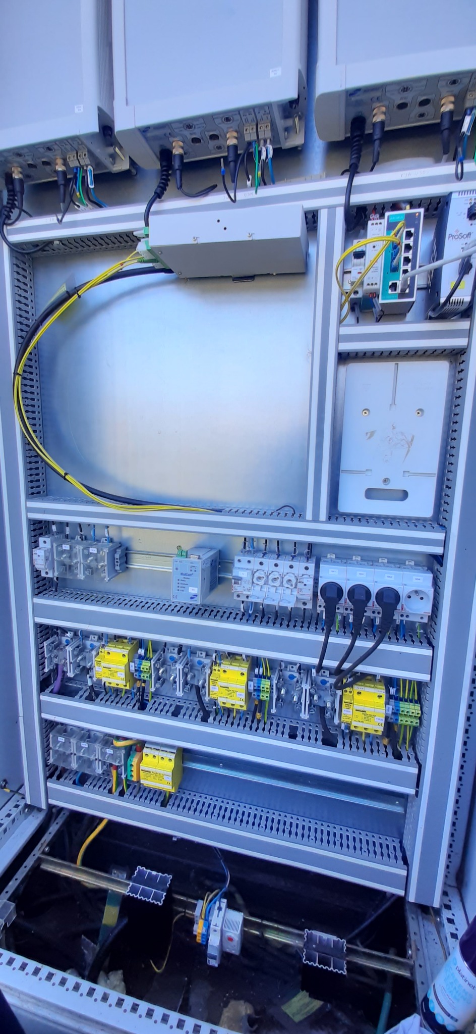 Otwarta szafa sterownicza z widocznymi elementami instalacji elektrycznej, w tym gniazdami, bezpiecznikami i przewodami w kolorach żółtym i czarnym, z elementami automatyki i ProSoft na boku.