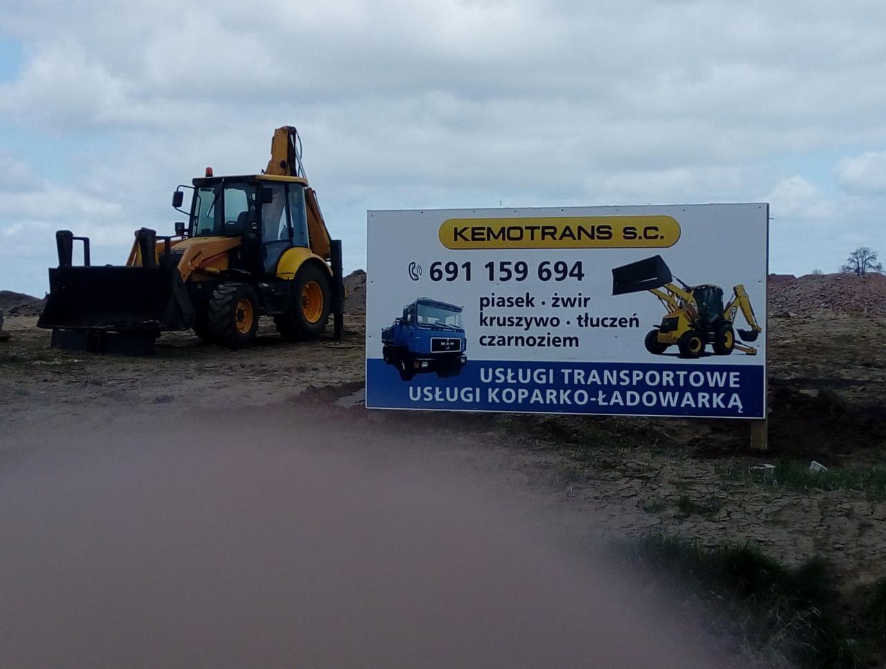 Żółto-czarna koparko-ładowarka na placu budowy, obok tablica reklamowa firmy Kemotrans S.C. oferującej usługi transportowe i koparko-ładowarką, z numerem telefonu i informacją o sprzedaży kruszyw.