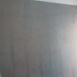 Ściana pokryta szarą, dekoracyjną gładzią z widoczną strukturą, drabina malarska oparta o ścianę, ciemne drzwi balkonowe w tle.