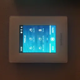 Biały panel sterowania rekuperacją Komfovent zamontowany na ścianie, wyświetlający aktualne parametry pracy systemu wentylacji: temperatury, wilgotność i przepływ powietrza.