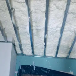 Poddasze z widoczną konstrukcją metalową wypełnioną gęstą, białą pianą izolacyjną, zabezpieczone folią malarską na ścianie, widok z dołu w trakcie prac.