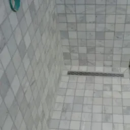 Wykończona kabina prysznicowa z jasnymi, kwadratowymi płytkami marmurowymi i odpływem liniowym. Widoczne przygotowane miejsce na baterię.