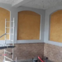 Ściana z dekoracyjną, złoto-żółtą fakturą wnęk, szara listwa, dolna część ściany wykończona imitacją cegły, widoczny fragment rusztowania i folii ochronnej.