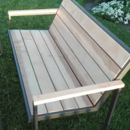 Nowoczesna ławka ogrodowa z drewnianym siedziskiem i metalową ramą, ustawiona na trawniku w słoneczny dzień. Widoczne detale łączeń drewna i metalu.