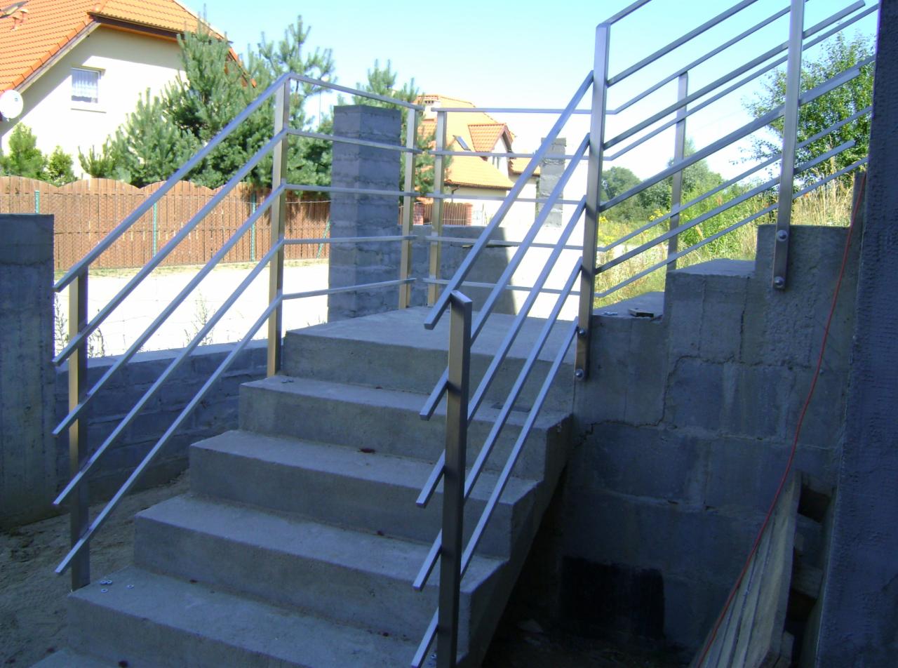 Betonowe schody zewnętrzne z prostą, metalową barierką na tle domów jednorodzinnych.