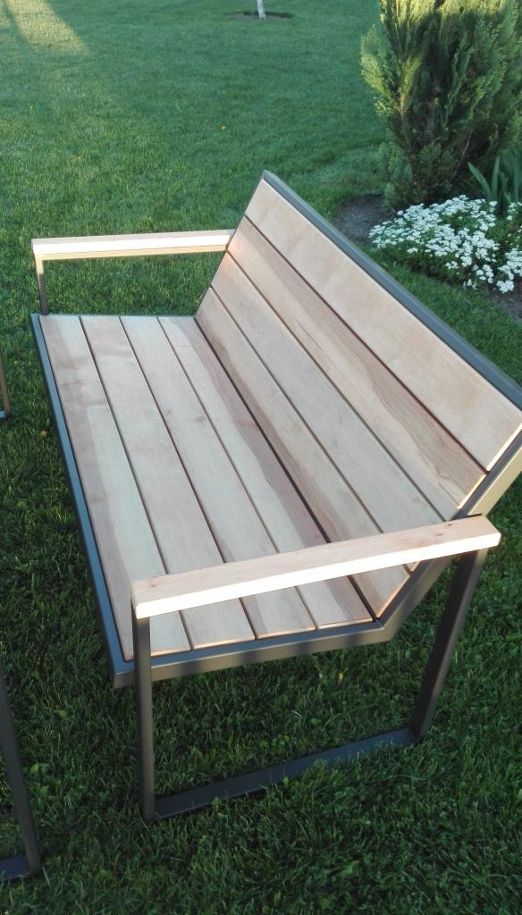 Nowoczesna ławka ogrodowa z drewnianym siedziskiem i metalową ramą, ustawiona na trawniku w słoneczny dzień. Widoczne detale łączeń drewna i metalu.
