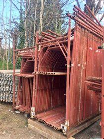 Stos czerwonych, metalowych ram rusztowań budowlanych, ustawionych pionowo, z widocznymi śladami użytkowania i rdzy, na tle drzew bez liści i stosu rur.