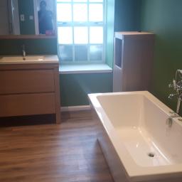 Adam's building - Nowoczesna łazienka z drewnianymi meblami, białą wanną i zielonymi ścianami, widok z perspektywy drzwi, w oknie odbija się sylwetka osoby.