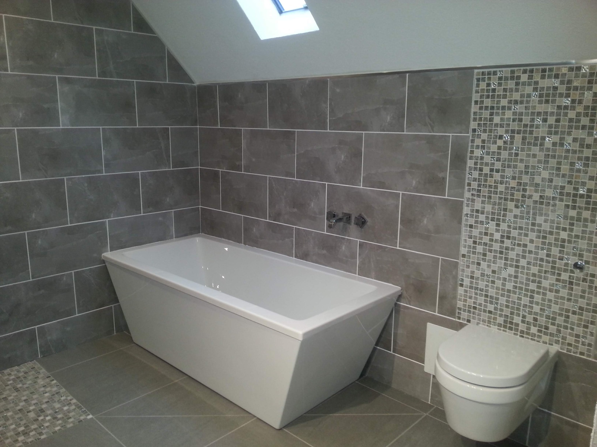 Nowoczesna łazienka z szarymi płytkami imitującymi kamień, mozaiką, białą wanną prostokątną i podwieszaną toaletą, oświetlona światłem z okna dachowego.