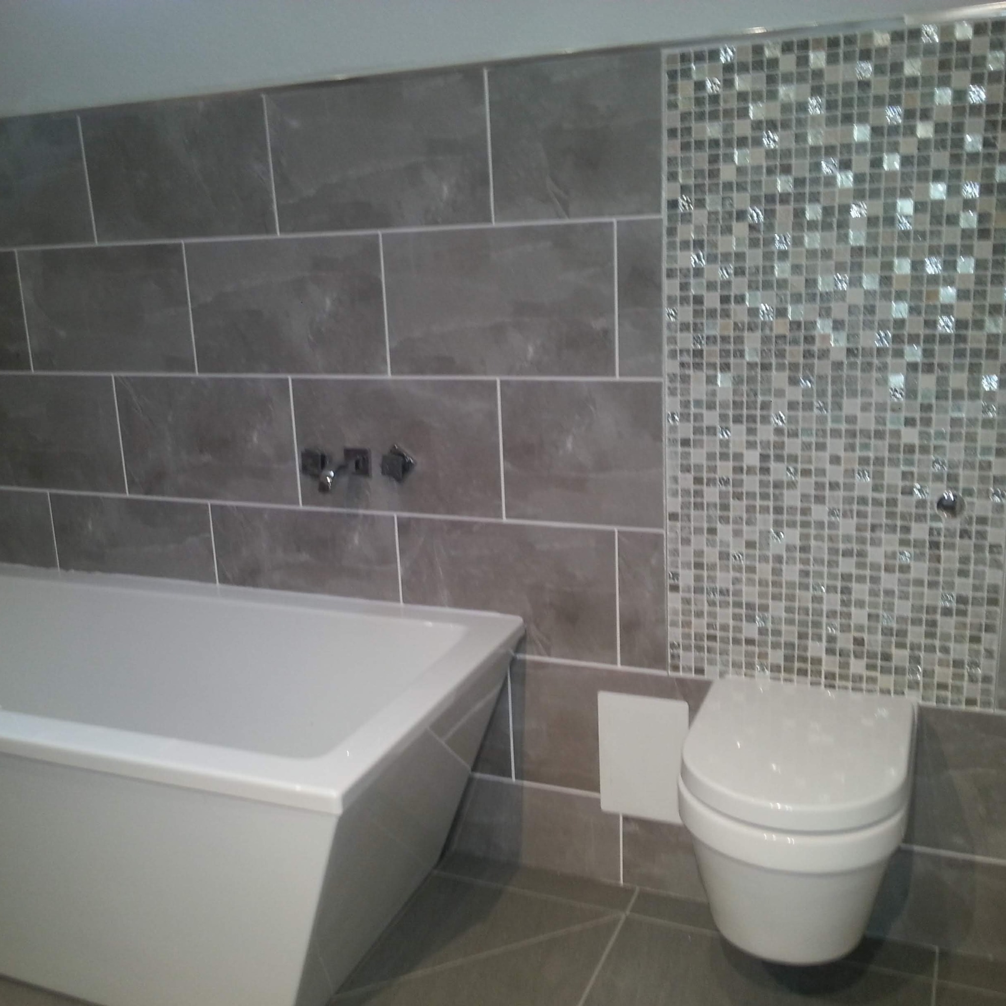 Wykończona łazienka z wanną, podwieszaną toaletą i szarymi płytkami imitującymi kamień, uzupełnionymi mozaiką z połyskującymi elementami.