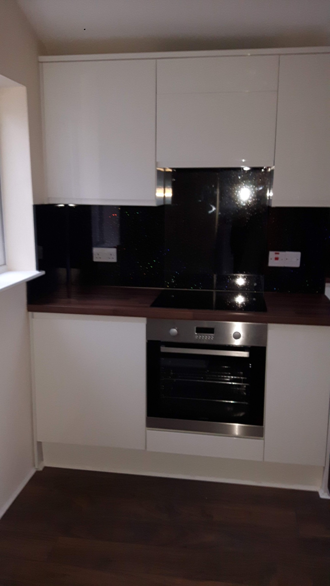 Nowoczesna kuchnia z białymi szafkami, ciemnym blatem i błyszczącym czarnym panelem nad indukcją, widoczne gniazdka elektryczne.