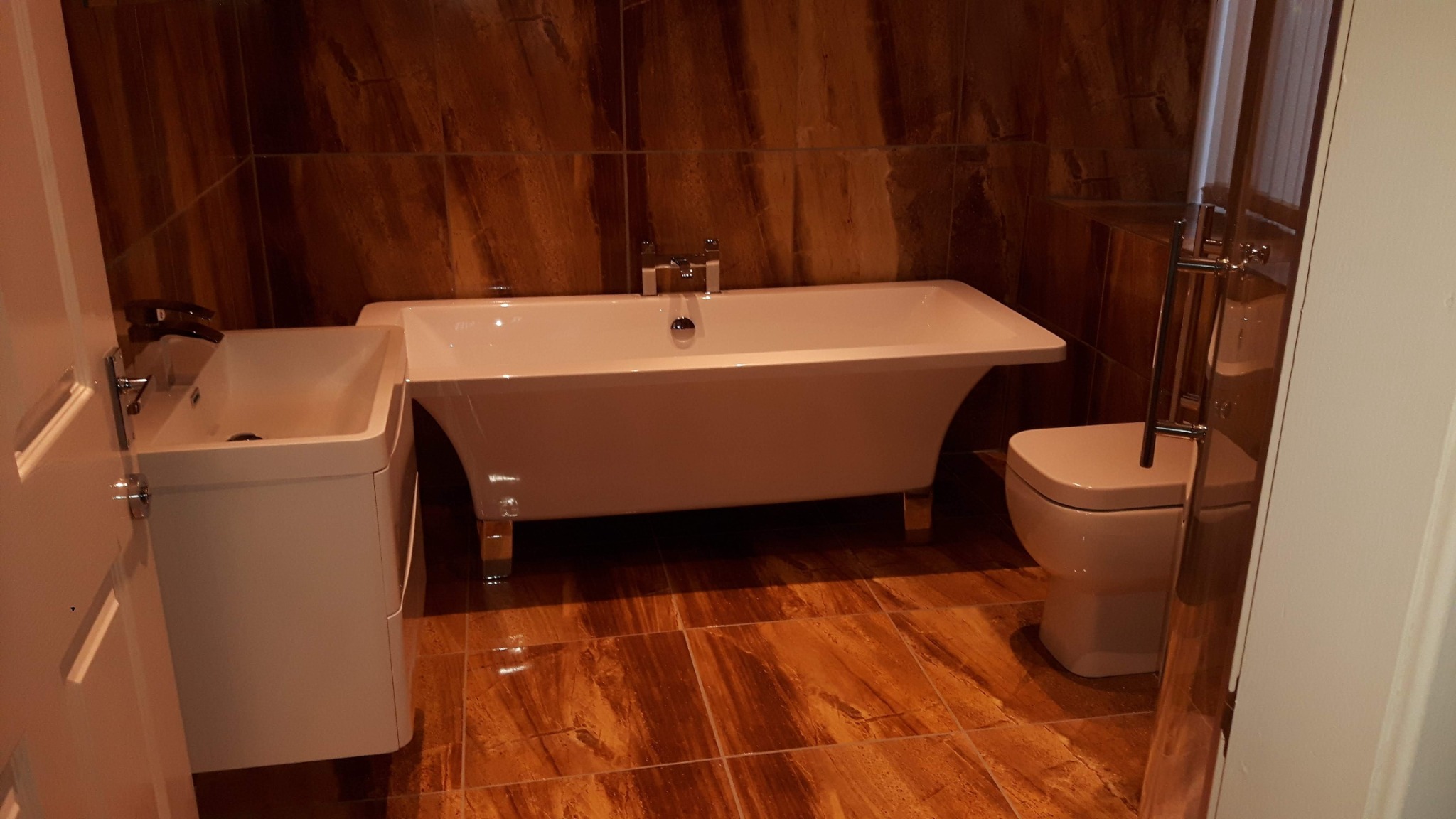 Nowoczesna łazienka z wolnostojącą wanną na złotych nóżkach, umywalką z szafką i toaletą, wykończona płytkami w odcieniach brązu i beżu.