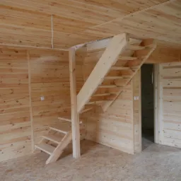 Wnętrze domu szkieletowego z drewnianą antresolą i otwartymi schodami, widoczne drewniane ściany i podłoga z płyty OSB, drzwi drewniane z przeszkleniami.