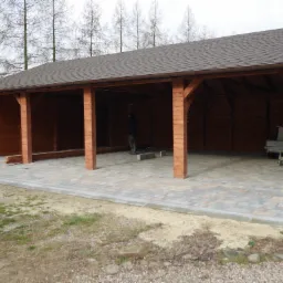 Zadaszona konstrukcja z drewna z brązowym gontem bitumicznym, podłoże wyłożone kostką brukową, w tle widoczne drzewa bez liści.