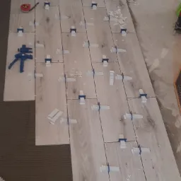 Układanie podłogi z płytek imitujących drewno, z użyciem systemu poziomowania płytek, widoczna miarka i zacisk stolarski.