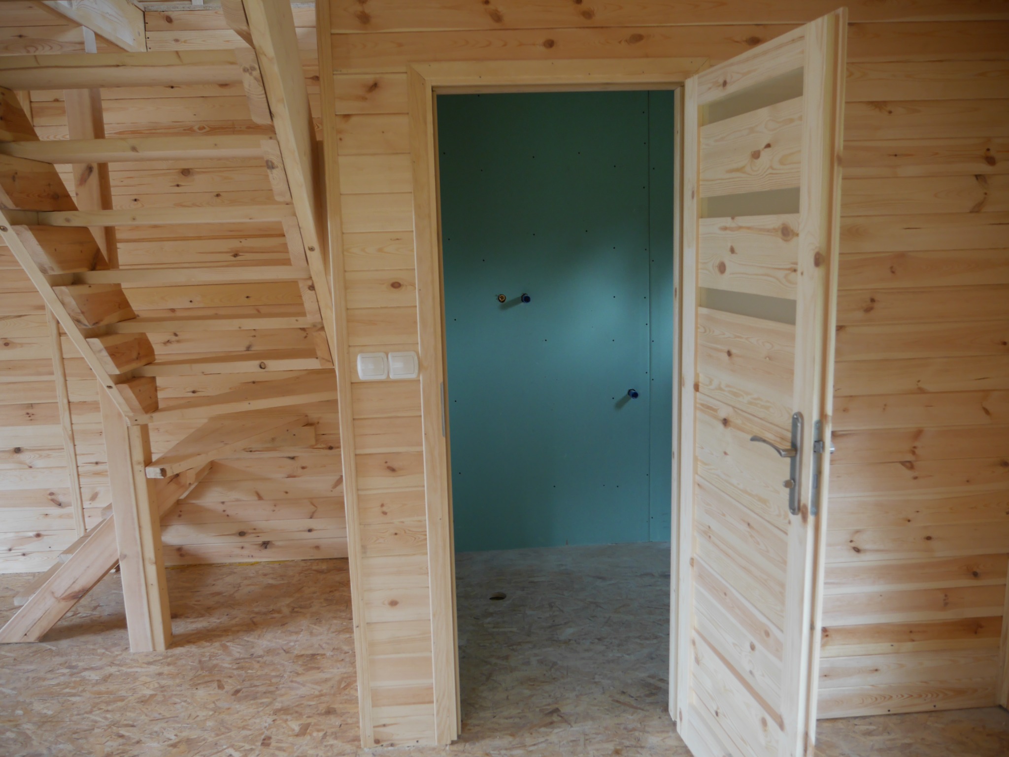 Surowe wnętrze domu w trakcie budowy, z drewnianymi ścianami, schodami i otwartymi drewnianymi drzwiami z przeszkleniami, ukazującymi przygotowaną pod instalacje ścianę w pomieszczeniu obok.