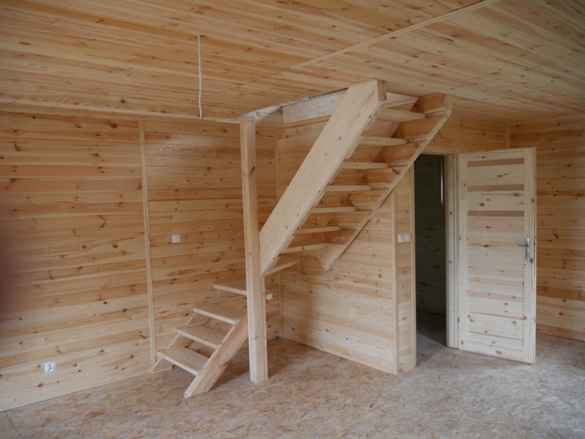 Wnętrze domu szkieletowego z drewnianą antresolą i otwartymi schodami, widoczne drewniane ściany i podłoga z płyty OSB, drzwi drewniane z przeszkleniami.