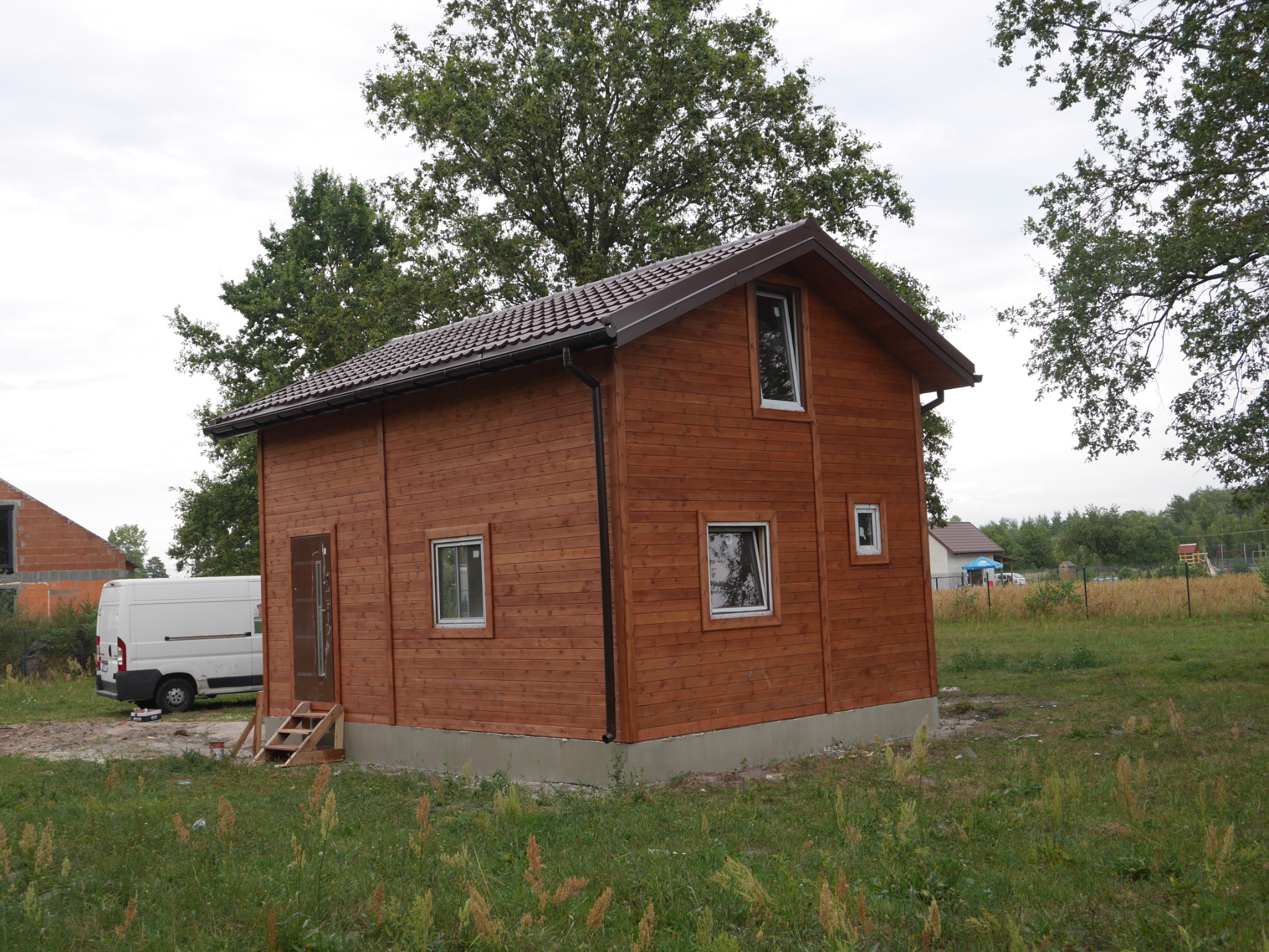 Nowo wybudowany, jednopiętrowy dom z brązowych desek z brązowym dachem, widoczny z boku, z prowizorycznymi schodami do drzwi wejściowych i białym samochodem dostawczym w tle.