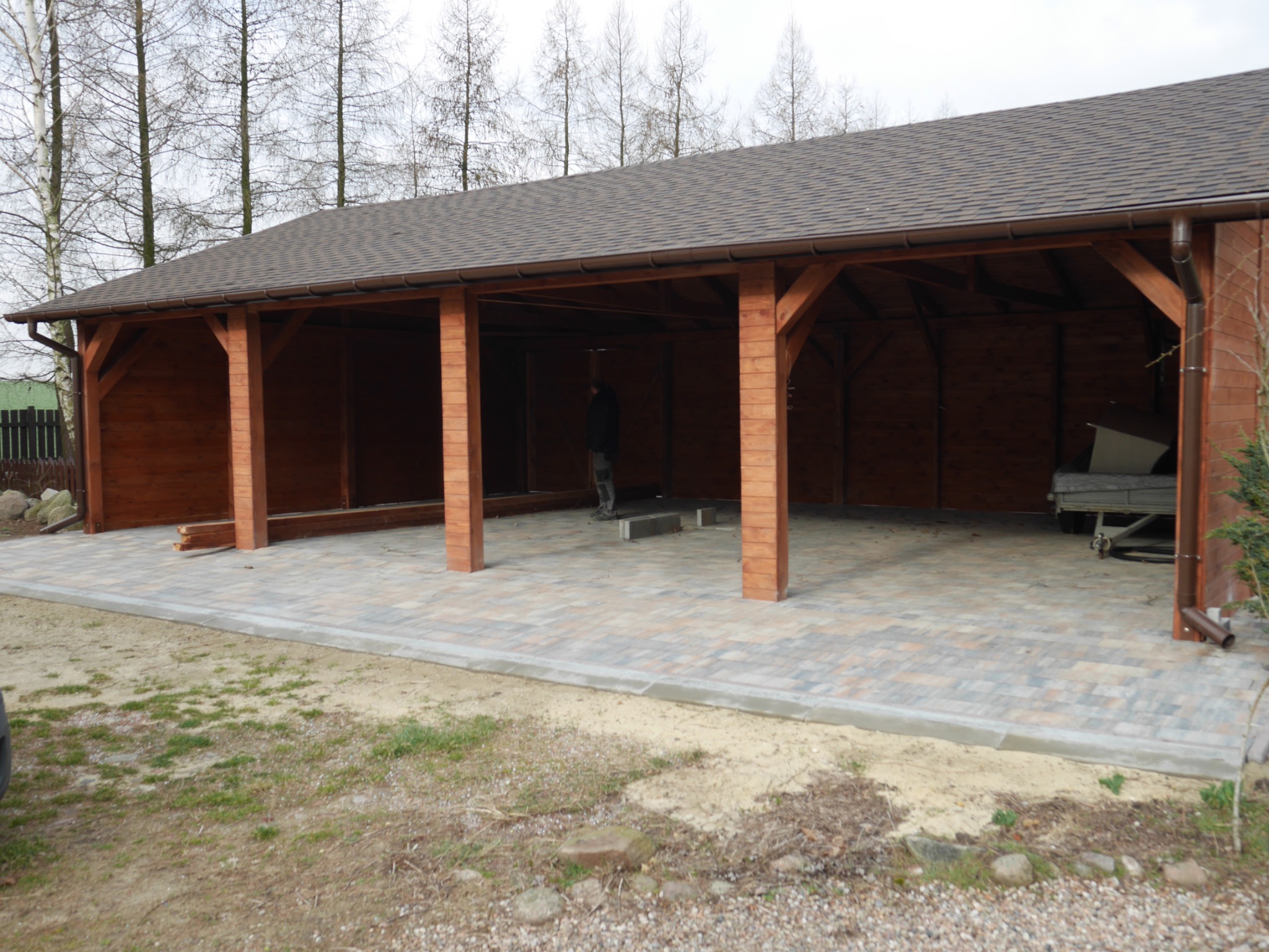 Zadaszona konstrukcja z drewna z brązowym gontem bitumicznym, podłoże wyłożone kostką brukową, w tle widoczne drzewa bez liści.
