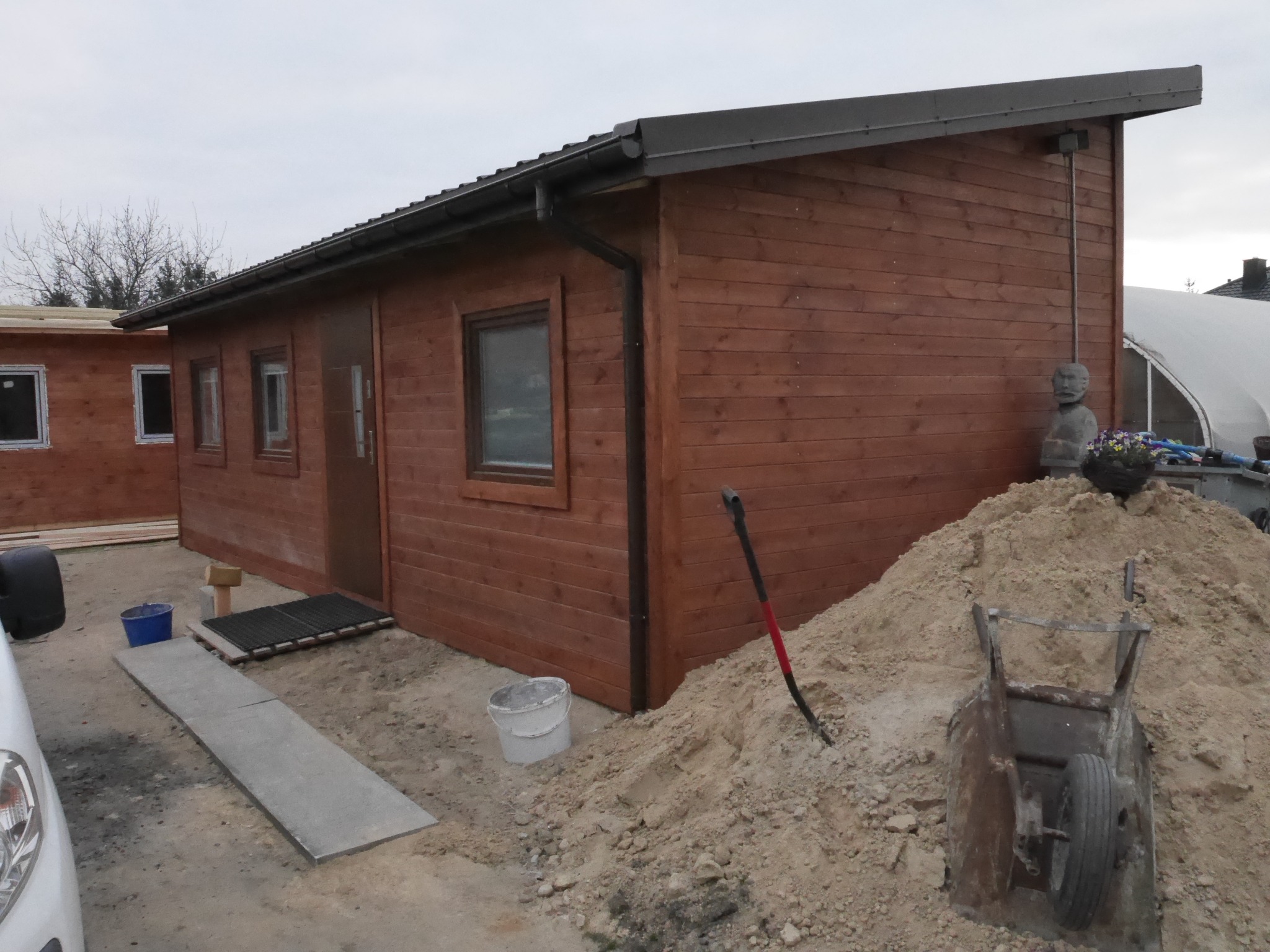 Nowoczesny, parterowy dom z drewna w trakcie budowy, z widocznym stosem piasku, taczką i narzędziami na placu budowy.