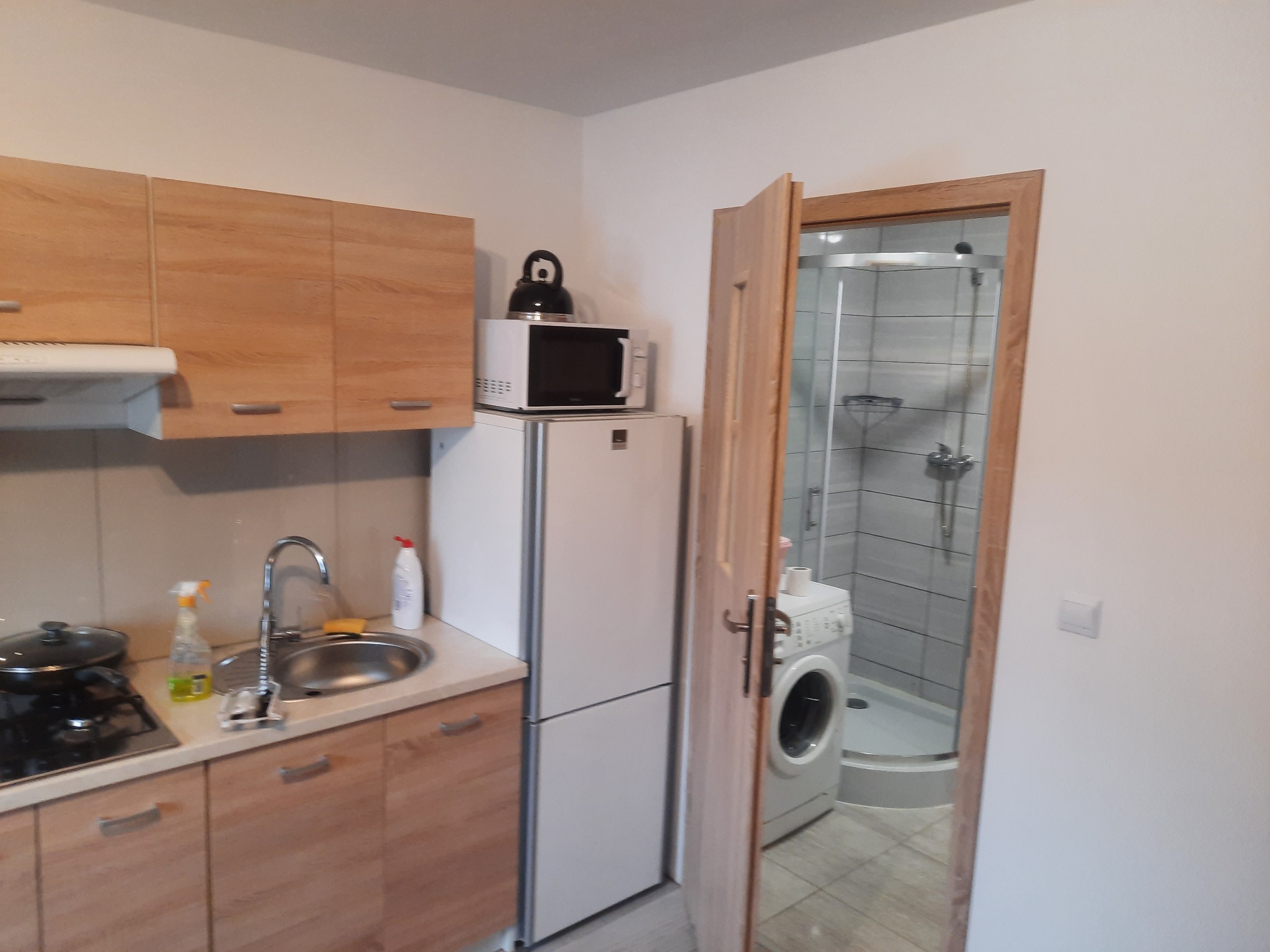 Niewielka kuchnia z drewnianymi szafkami i widokiem na łazienkę z kabiną prysznicową oraz pralką za otwartymi drzwiami