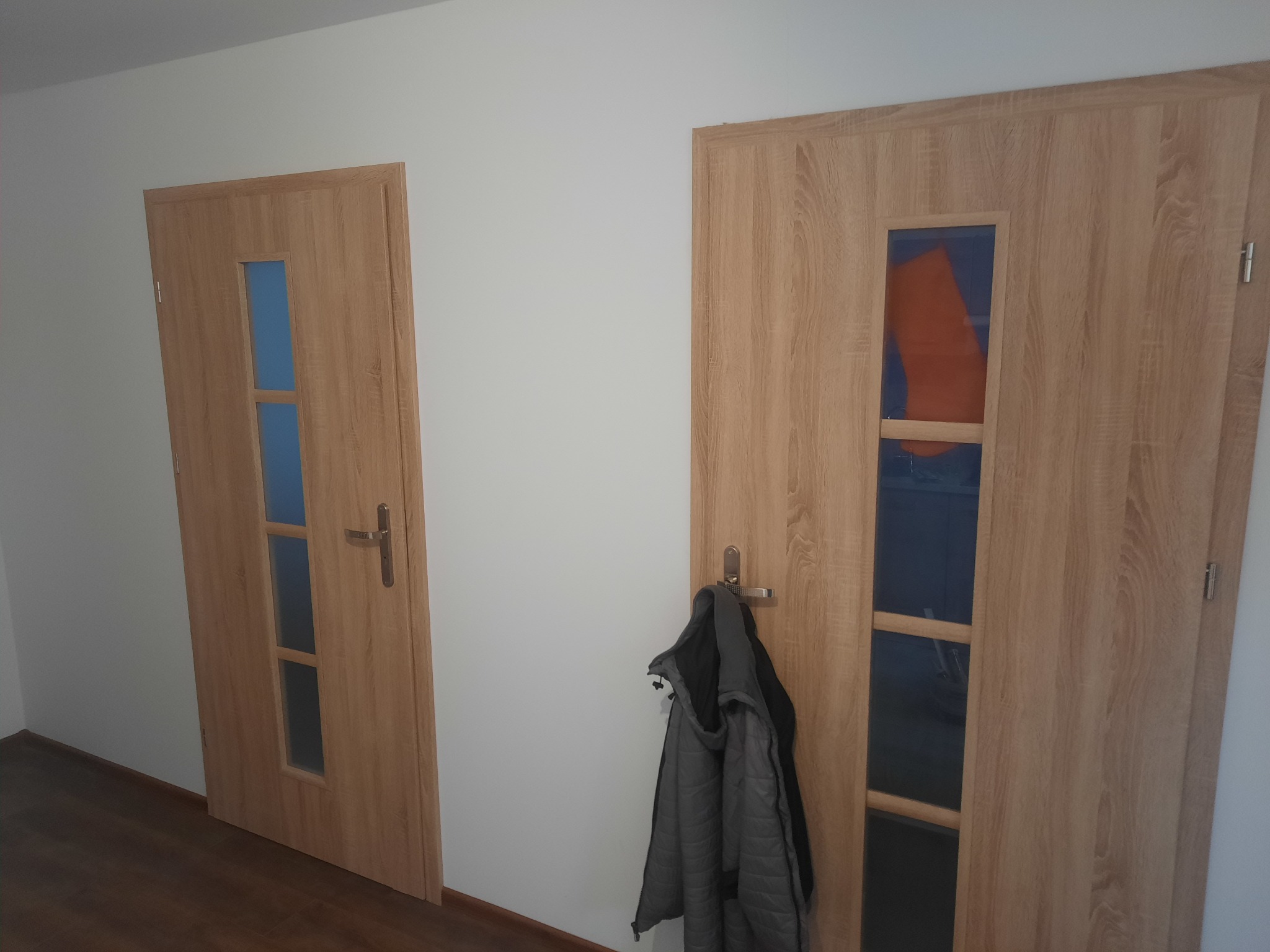Dwie wewnętrzne drzwi z drewnopodobnym fornirem i szybami; na klamce jednej z nich wisi szara kurtka.