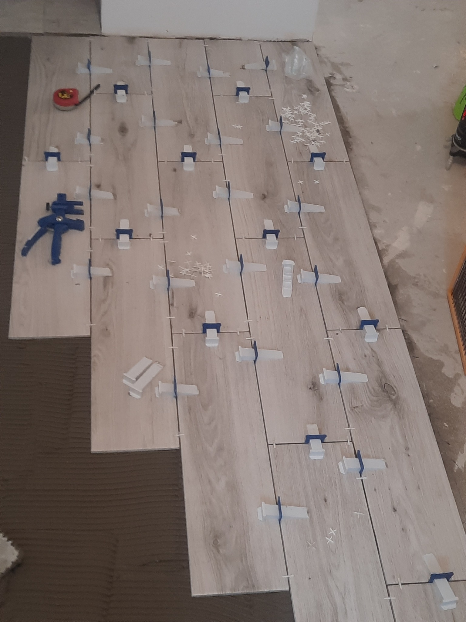 Układanie podłogi z płytek imitujących drewno, z użyciem systemu poziomowania płytek, widoczna miarka i zacisk stolarski.