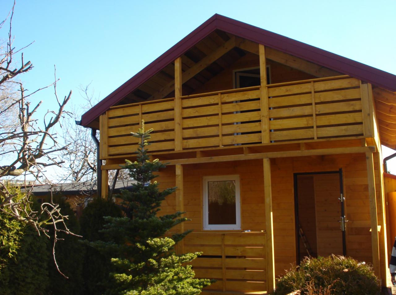 Drewniany domek letniskowy z balkonem, jasne drewno, czerwony dach, małe okno, otoczony zielenią i drzewami bez liści, błękitne niebo.