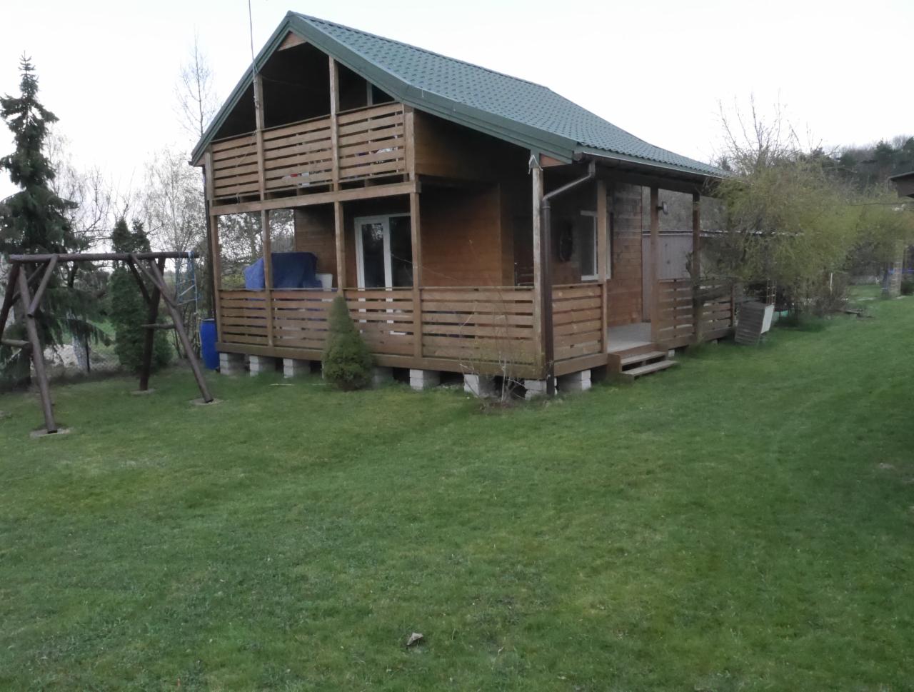 Dwukondygnacyjny drewniany domek letniskowy z zielonym dachem, drewnianymi balustradami na balkonach i werandzie, posadowiony na betonowych bloczkach, otoczony trawnikiem i drzewami.