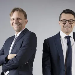 Portret dwóch uśmiechniętych prawników w granatowych garniturach na jasnoszarym tle, jeden z założonymi rękami.