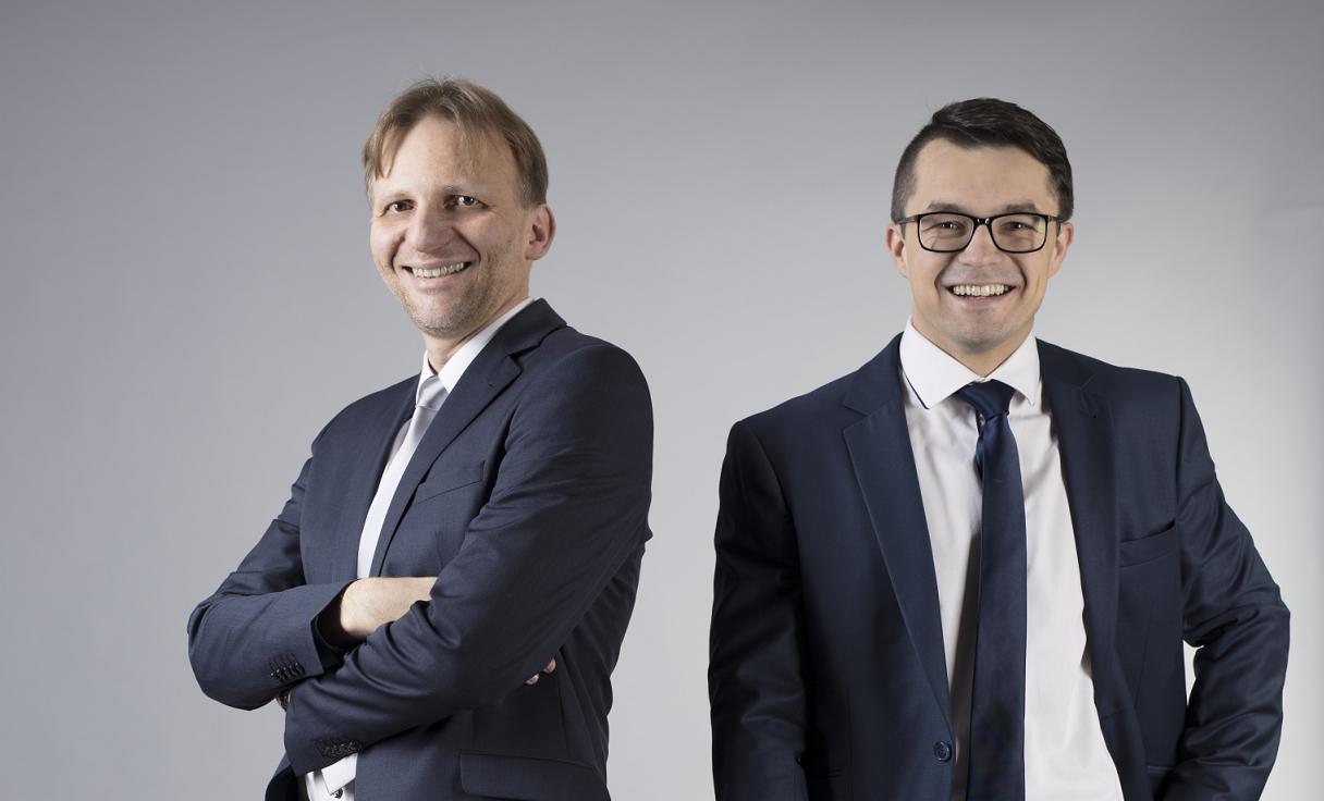 Portret dwóch uśmiechniętych prawników w granatowych garniturach na jasnoszarym tle, jeden z założonymi rękami.