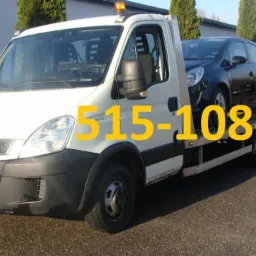 Biała laweta Iveco z czarnym samochodem osobowym na platformie, zaparkowana na asfaltowej drodze, w tle rząd wysokich, zielonych drzew.