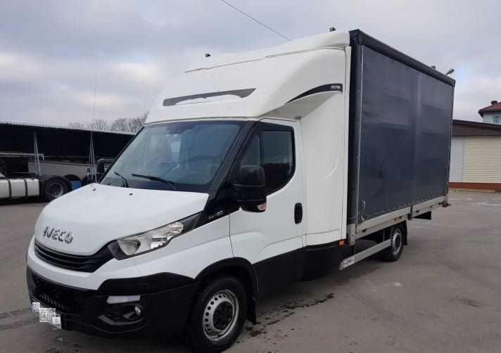 Biała ciężarówka Iveco z plandeką na parkingu, widok z przodu i boku, pochmurne niebo w tle.