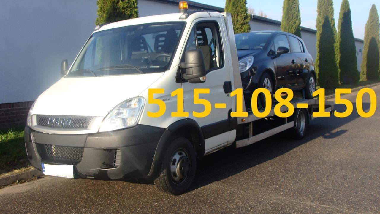 Biała laweta Iveco z czarnym samochodem osobowym na platformie, zaparkowana na asfaltowej drodze, w tle rząd wysokich, zielonych drzew.