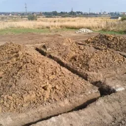 Wykopane rowy pod fundamenty z pryzmami ziemi, widok na odległe zabudowania wiejskie i domy jednorodzinne.