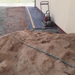 Perfect Home - Układanie kostki brukowej przy budynku: zharmonizowane szare kostki z żółtym akcentem, zagęszczarka spalinowa Honda obok, widoczny kopiec piasku z zielonym wężem.