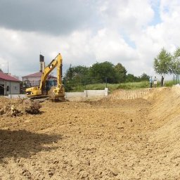 Perfect Home - Żółta koparka wyrównuje teren pod budowę, widoczne fundamenty i palisada zabezpieczająca skarpę, w tle zabudowania wiejskie