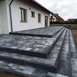 Ułożona kostka brukowa na tarasie i schodach przed domem w Czerwionce-Leszczynach. Widoczne szare i czarne kostki tworzące wzór na powierzchni.