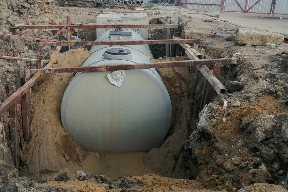 Dwa zbiorniki bezodpływowe zakopane w wykopie, zabezpieczone drewnianą konstrukcją, widoczne rury i piasek.