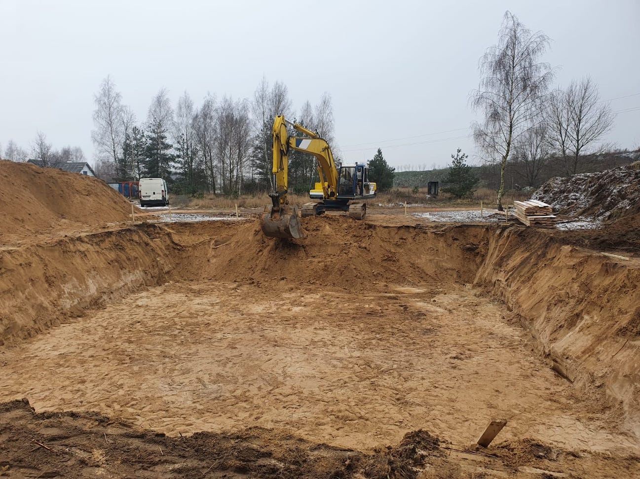 Żółta koparka w trakcie prac ziemnych związanych z wykopem pod fundamenty, widoczne deski szalunkowe i biały samochód dostawczy w tle.