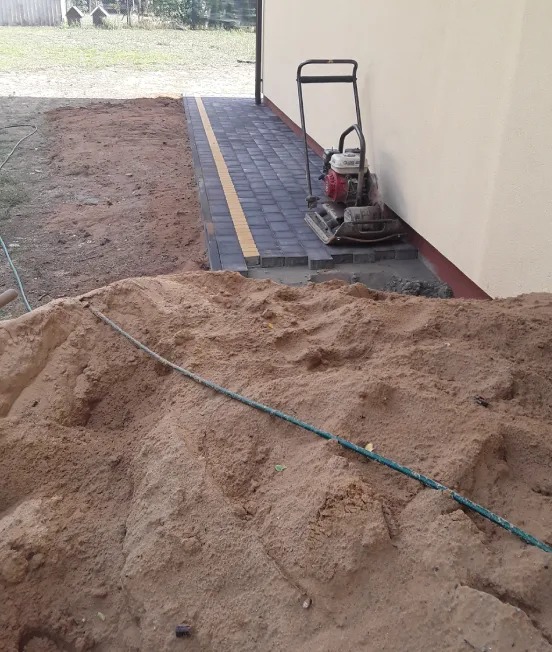 Układanie kostki brukowej przy budynku: zharmonizowane szare kostki z żółtym akcentem, zagęszczarka spalinowa Honda obok, widoczny kopiec piasku z zielonym wężem.