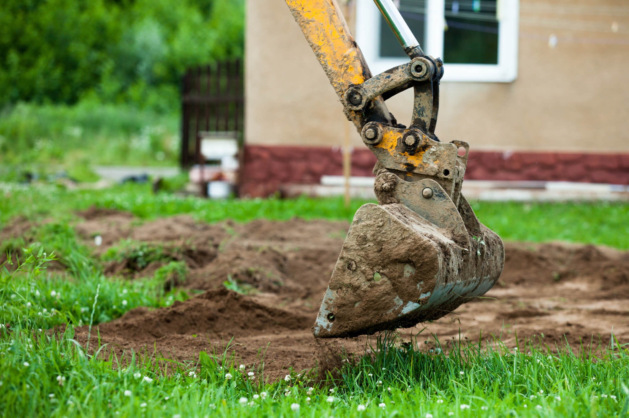 Żółta koparka z łyżką pełną ziemi, przygotowująca teren pod budowę, widoczny fragment budynku w tle.