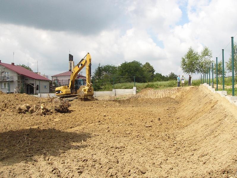 Żółta koparka wyrównuje teren pod budowę, widoczne fundamenty i palisada zabezpieczająca skarpę, w tle zabudowania wiejskie
