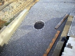 Ułożona z szarej kostki granitowej aleja w kształcie wachlarza z okrągłą studzienką kanalizacyjną pośrodku, widoczne deski i świeżo wylany beton.