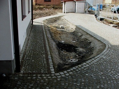 Fragment podjazdu z kostki granitowej z przygotowanym korytem na odprowadzenie wody deszczowej wzdłuż ściany budynku.